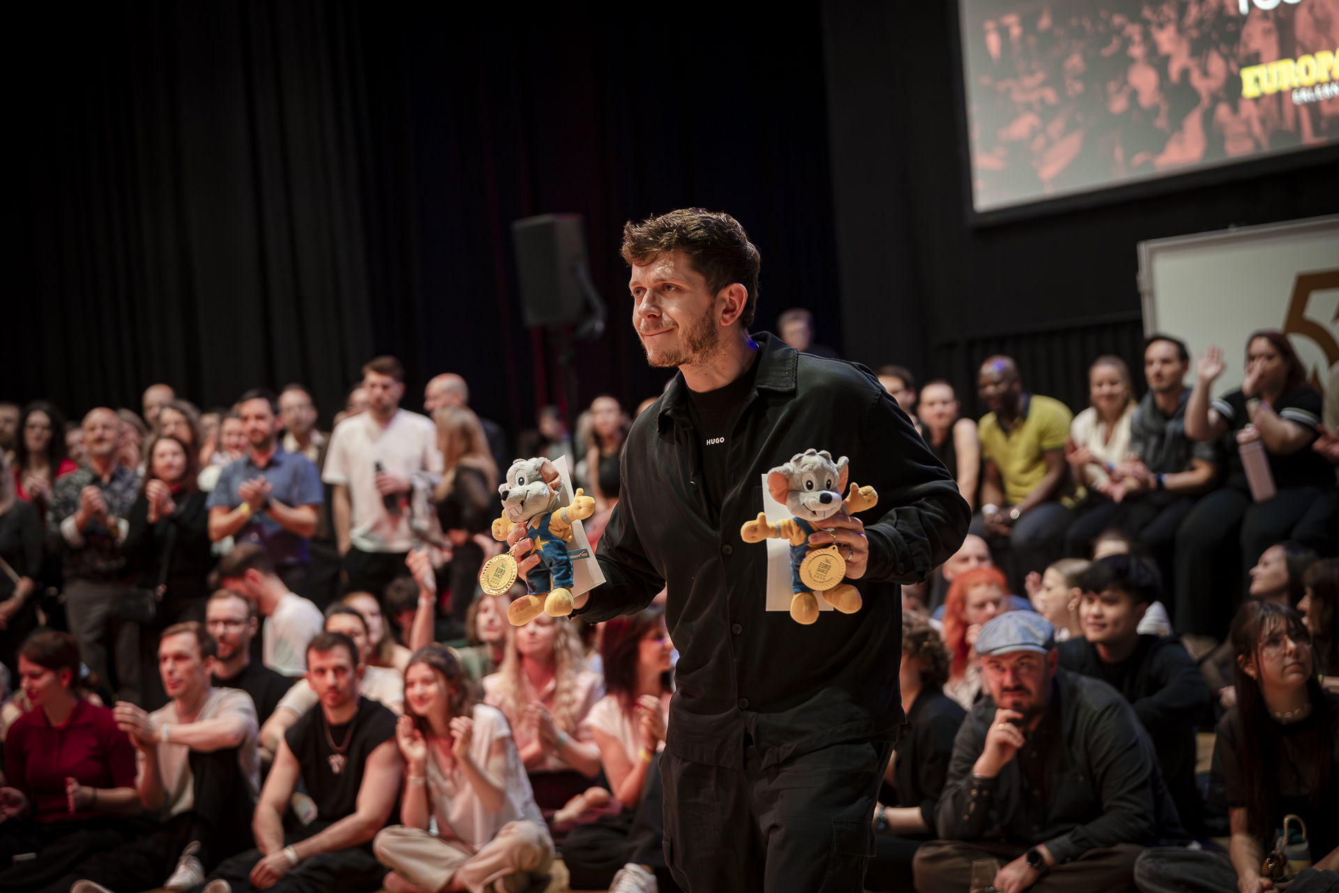 ThiMaus A man holding two stuffed toy mouse figures, standing in front of an audience